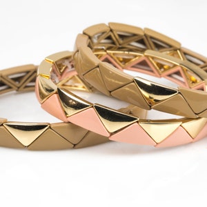 Puede incluir: Tres pulseras apilables con un diseño triangular. Las pulseras están hechas de un material beige y rosa con detalles dorados.