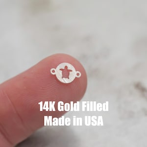 May include: A close-up of a 14K gold filled turtle charm. The charm is round and has a cutout of a turtle in the center. The charm is made in the USA.