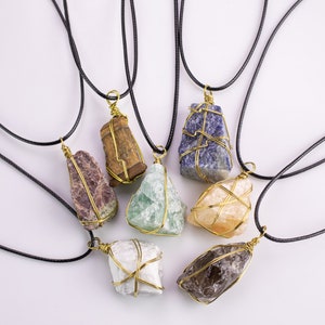 May include: A collection of gemstone pendants on black cords. Each raw stone is encased in gold-coloured wire. The stones are various colours, including purple, green, blue, and brown. The pendants are displayed against a white backdrop.