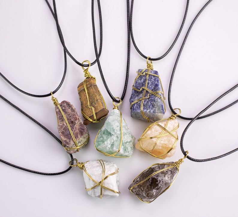 May include: A collection of gemstone pendants on black cords. Each raw stone is encased in gold-coloured wire. The stones are various colours, including purple, green, blue, and brown. The pendants are displayed against a white backdrop.