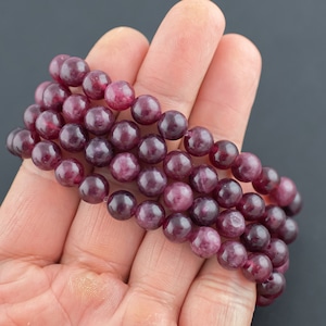 May include: A set of three beaded bracelets with round, polished, dark pink beads. The bracelets are held in a hand against a dark gray background. The beads have a smooth, glossy finish, and the color is a deep, rich shade of pink.