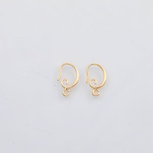 May include: Two gold-colored earring hooks with a decorative swirl design. The hooks have a curved shape with a small loop at the bottom for attaching charms or beads. The earrings are set against a white background.