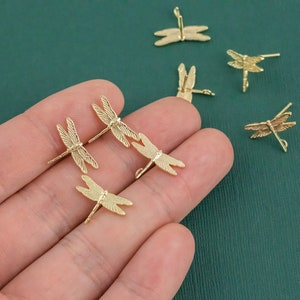 Op de afbeelding: Een hand die een aantal gouden libellenbedels vasthoudt. De bedels zijn gedetailleerd en hebben een delicaat ontwerp.