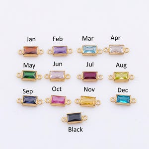 Könnte beinhalten: Goldfarbene, rechteckige Charms mit farbigen Edelsteinen, die jeweils einen Monat von Januar bis Dezember darstellen, sowie ein schwarzer Charm. Die Monate sind über jedem Charm beschriftet. Diese Charms sind für Schmuck.