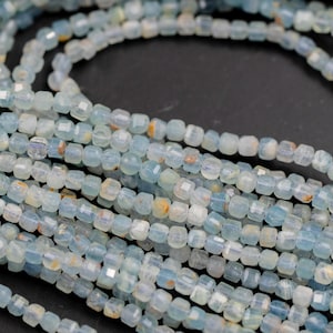 Może przedstawiać: Bliska perspektywa sznurów małych, jasnoniebieskich koralików z kamieni szlachetnych. Koraliki są kwadratowe i fasetowane, tworząc efekt iskrzenia. Koraliki mają lekko przezroczystą jakość, z nutami żółtego i białego. Koraliki są nawleczone, tworząc naszyjnik lub bransoletkę.