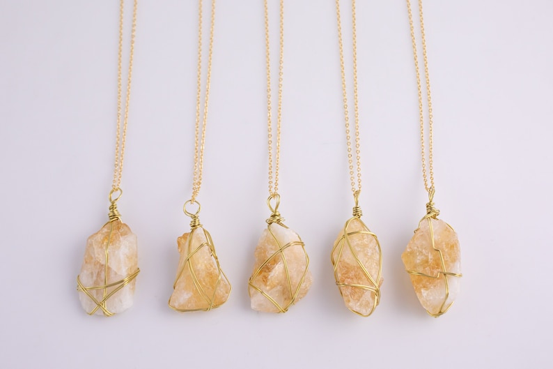 May include: Five necklaces featuring raw, light orange citrine crystals, each encased in gold-coloured wire and suspended from fine gold chains. The crystals' shapes and sizes differ, offering a natural look. The background is plain white.