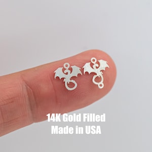 May include: Two silver dragon charms with a loop at the top. The charms are on a finger and the text "14K Gold Filled Made in USA" is below the charms.