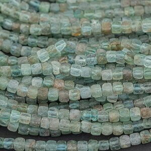 May include: Close-up of strands of square-shaped beads in shades of blue-green and brown. The beads are strung together, creating a textured, organic pattern. The beads are likely used for jewelry making.