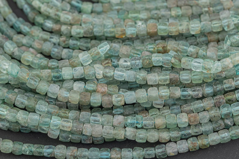 May include: Close-up of strands of square-shaped beads in shades of blue-green and brown. The beads are strung together, creating a textured, organic pattern. The beads are likely used for jewelry making.