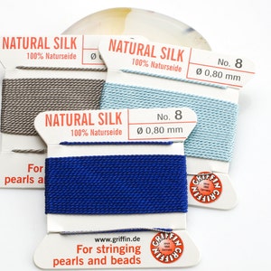 Pu&ograve; includere: Tre bobine di filo di seta naturale per infilare perle e perline. Le bobine sono etichettate "Natural Silk 100% Naturseide" e "No. 8 &Oslash; 0,80 mm". I colori del filo sono grigio, azzurro e blu scuro.