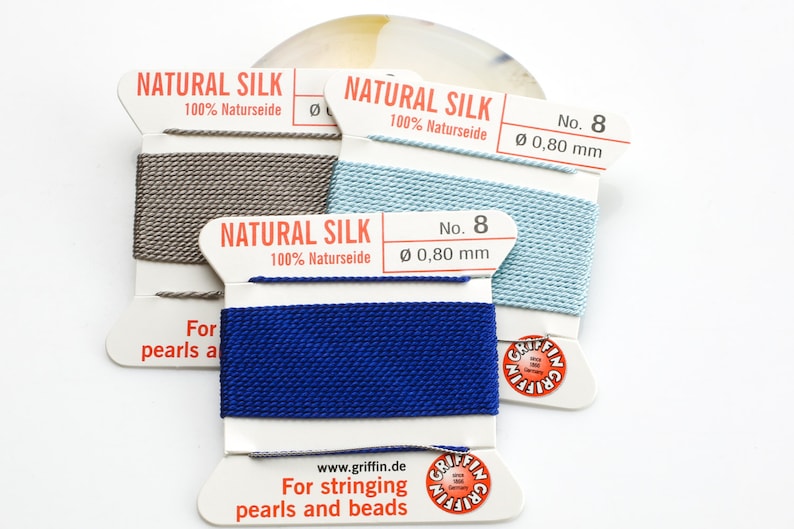 Pu&ograve; includere: Tre bobine di filo di seta naturale per infilare perle e perline. Le bobine sono etichettate "Natural Silk 100% Naturseide" e "No. 8 &Oslash; 0,80 mm". I colori del filo sono grigio, azzurro e blu scuro.
