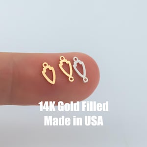 Puede incluir: Tres pequeños dijes dorados en forma de escudo con un lazo en la parte superior para hacer joyas. Un dije es dorado, uno plateado y otro relleno de oro. El texto de la imagen dice "14K Gold Filled Made in USA."