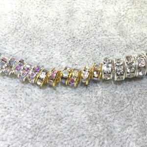 May include: A strand of cylindrical beads featuring embedded clear crystals. The beads on the left side are set in gold-toned metal with iridescent crystal reflections, while the beads on the right side are set in silver-toned metal with clear crystals.