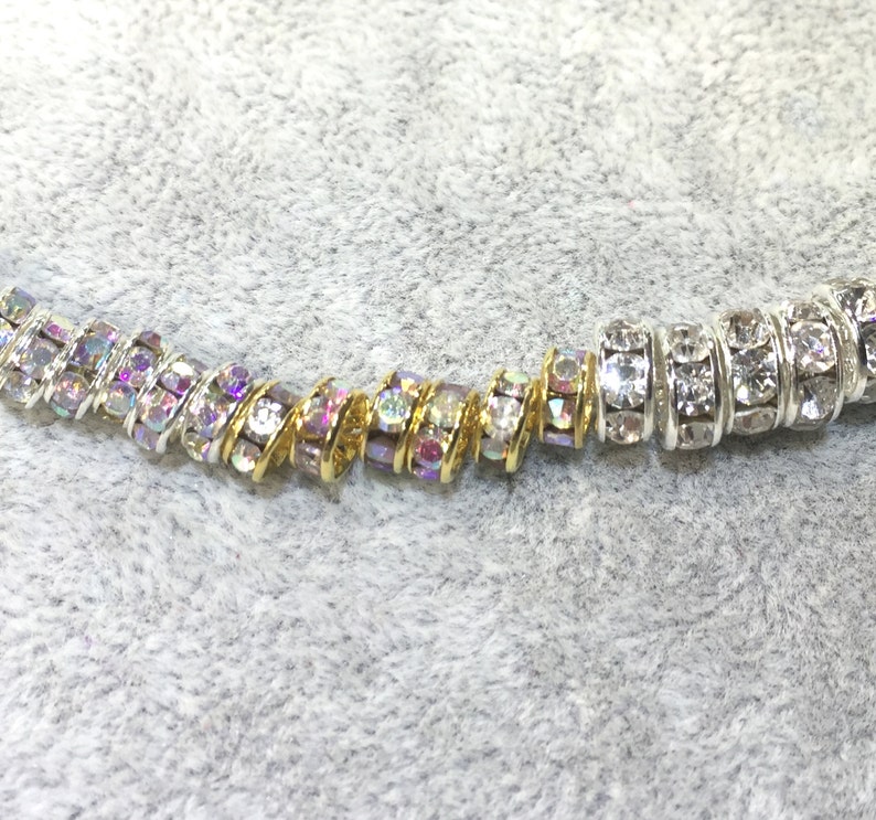 May include: A strand of cylindrical beads featuring embedded clear crystals. The beads on the left side are set in gold-toned metal with iridescent crystal reflections, while the beads on the right side are set in silver-toned metal with clear crystals.