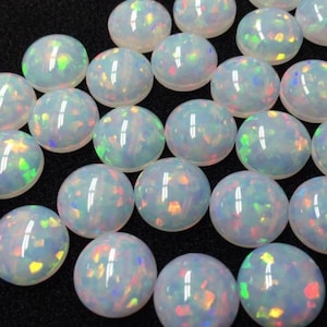 Può includere: Una collezione di cabochon di opale bianco con riflessi arcobaleno iridescenti. I cabochon sono rotondi e lisci e sono disposti in uno schema casuale.