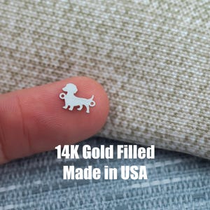 以下が含まれることがあります： ジュエリーに取り付けるためのループが付いた、小さなシルバー色のダックスフンドチャーム。チャームは指の上に表示され、その下に白いブロック文字で「14K Gold Filled Made in USA」というテキストが表示されています。