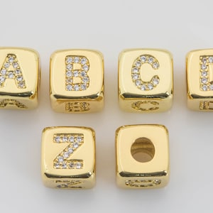 May include: Gold-toned alphabet letter beads with clear cubic zirconia stones. The beads are in the shape of a cube and feature the letters A, B, C, D, and Z. There is also a plain gold bead with a hole in the center.