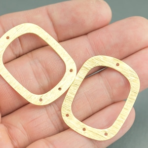 May include: Two gold-colored metal frames with rounded corners and four holes each. The frames are slightly different sizes and are lying on a hand.