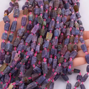 May include: Strands of tumbled, rectangular gemstones in shades of purple, blue, and brown are strung together with pink string and small, clear beads. The gemstones are held in a hand, showcasing the color variations and textures of the stones.