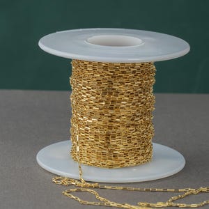 May include: A spool of gold-colored chain, wound around a white plastic spool. The chain is made of small, rectangular links.