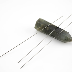 May include: Three silver needles resting on a green and gray stone. The needles are long and thin, and the stone is smooth and polished.