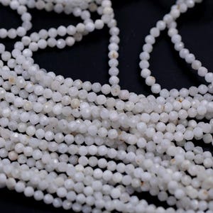 May include: Close-up of multiple strands of white, faceted beads. The beads are small and round, with a slightly iridescent sheen. The strands are arranged in a flowing, layered pattern against a dark background, suggesting jewelry making supplies.
