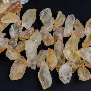 May include: A strand of raw, natural, light yellow citrine gemstone beads. The beads are irregular in shape and have a translucent, glassy appearance. The beads are strung on a thin, orange thread.