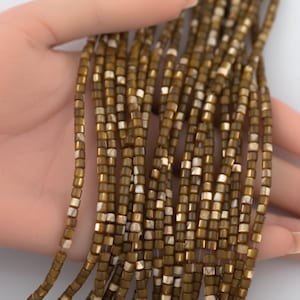 May include: Multiple strands of small, square-shaped beads in a metallic bronze and iridescent cream color palette. The beads are strung together in long, uniform lines, showcasing a reflective, polished finish with subtle variations in tone.