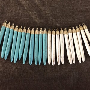 May include: A set of 18 pendant charms, half are turquoise and half are white. The charms are long and slender with a gold-coloured cap at the top.