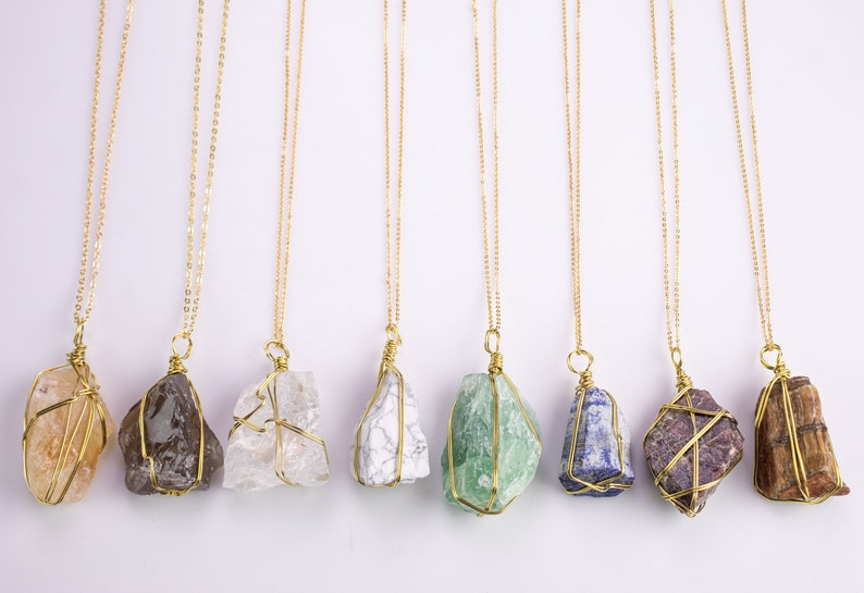 May include: A selection of gemstone necklaces. Each necklace has a unique, irregularly shaped stone wrapped in gold-coloured wire and suspended from a delicate gold-coloured chain. The stones vary in colour, including shades of brown, white, green, and purple.
