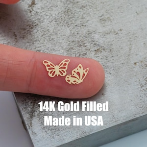 May include: Two small, gold-colored butterfly charms. The charms are cut out with intricate wing details. The text "14K Gold Filled Made in USA" is printed in white below the charms.