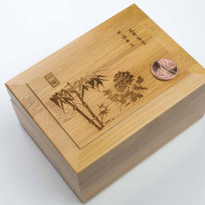May include: A wooden box with a hinged lid. The lid is decorated with an engraved design of bamboo and flowers. The box is made of light brown wood and has a natural finish. The box is closed and a copper coin is sitting on top of the lid.