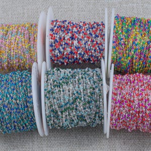 May include: Six spools of colorful beaded chain. The spools display a variety of colors, including yellow, pink, blue, green, and red. Each chain is made of small, round beads strung together. The spools are white and arranged in a row.
