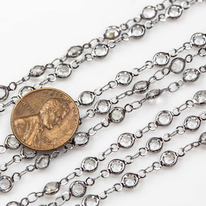 May include: A close-up of a silver chain with clear crystal beads. The chain is laid out in a straight line, with a penny placed in the center for scale.