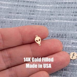 Può includere: Un piccolo ciondolo dorato a forma di alieno con due ritagli a forma di occhio e due piccoli anelli. Il testo "14K Gold Filled Made in USA" è visibile sotto.