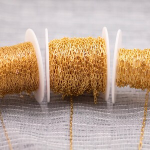 May include: Three spools of gold-colored chain are displayed against a gray background. The chain is made of small, interconnected links, and each spool is mounted on a white plastic holder. The chain is a bright, shiny gold color.