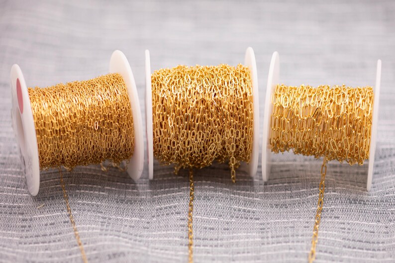 May include: Three spools of gold-colored chain are displayed against a gray background. The chain is made of small, interconnected links, and each spool is mounted on a white plastic holder. The chain is a bright, shiny gold color.