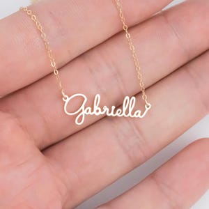 May include: A gold chain necklace with a delicate cursive nameplate pendant that reads "Gabriella".