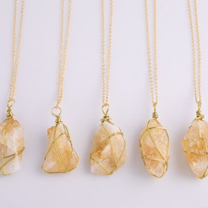 May include: Five gold necklaces, each featuring a raw, light orange citrine crystal pendant. The crystals are encased in gold wire and hang from fine gold chains. The backdrop is a plain white surface.