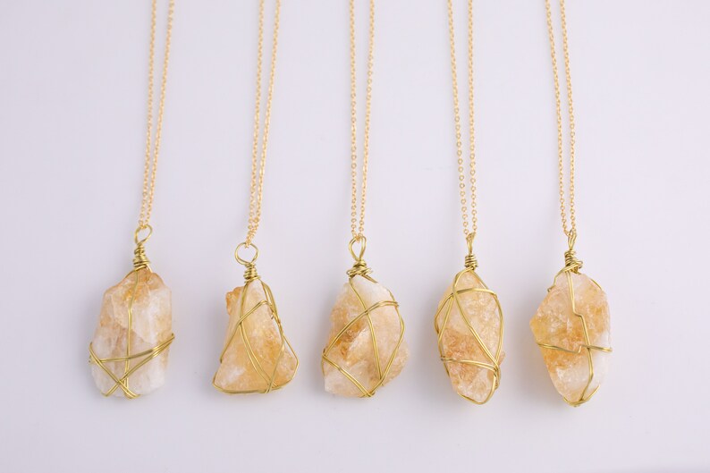 May include: Five gold necklaces, each featuring a raw, light orange citrine crystal pendant. The crystals are encased in gold wire and hang from fine gold chains. The backdrop is a plain white surface.