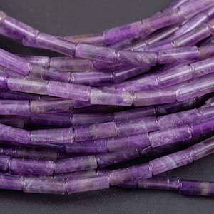 May include: Close-up of a collection of amethyst gemstone beads. The beads are cylindrical with a slightly rectangular shape and a polished finish. The color is a deep purple with some translucent areas.