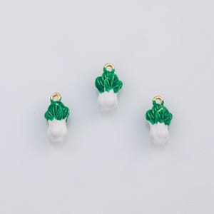Puede incluir: Tres dijes de bok choy en miniatura. Cada dije presenta una base blanca con una parte superior verde y frondosa y un lazo dorado para sujetarlo a joyas u otras manualidades. Los dijes están sobre un fondo blanco liso.