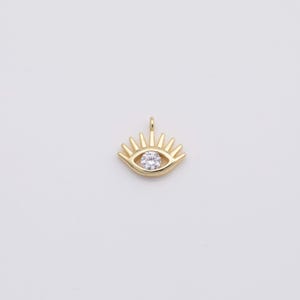 May include: Gold-colored eye pendant with a clear, round gemstone center. The pendant has a sunburst design around the eye, with a small loop at the top for a chain. The pendant is approximately 0.5 inches wide.