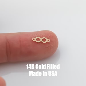 May include: A gold-filled infinity symbol charm with two small loops on either side. The charm is shown on a finger, and the text "14K Gold Filled Made in USA" is below the charm.
