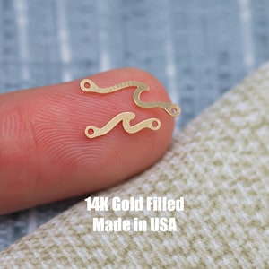 May include: Two gold-filled wave charms, one with a single curve and the other with two curves. The charms are on a finger, and the text "14K Gold Filled Made in USA" is below the charms.