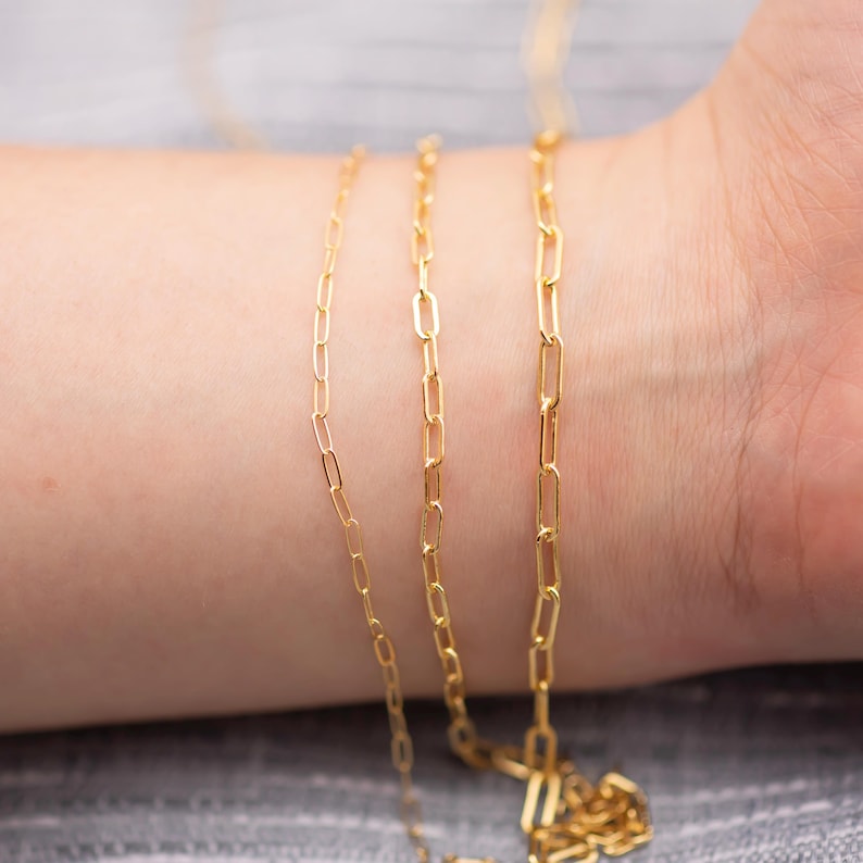 May include: Close-up of three delicate gold-colored chain necklaces. The chains have a rectangular link design. The necklaces are draped over a person's arm, with the background out of focus.