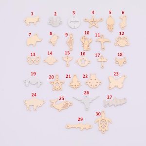 Puede incluir: Una colección de pequeños dijes de metal dorado en varias formas, incluyendo animales, criaturas marinas y diseños geométricos. Los dijes están numerados del 1 al 30 y se muestran sobre una superficie clara.