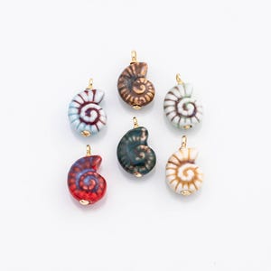 May include: A collection of six ceramic seashell pendants in various colors, including blue, brown, green, red, and white. Each pendant features a spiral design and a gold-colored bail for attaching to a necklace or bracelet. The pendants are displayed on a white background.