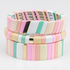 May include: Three stacked bracelets with colorful, rectangular beads in shades of pink, green, white, and beige. The bracelets have a gold accent.