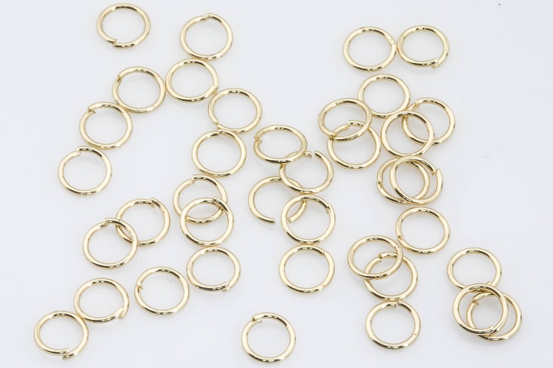 May include: A large quantity of gold jump rings, a type of jewellery finding, are scattered on a white surface. The rings are all the same size and are made of a shiny metal.